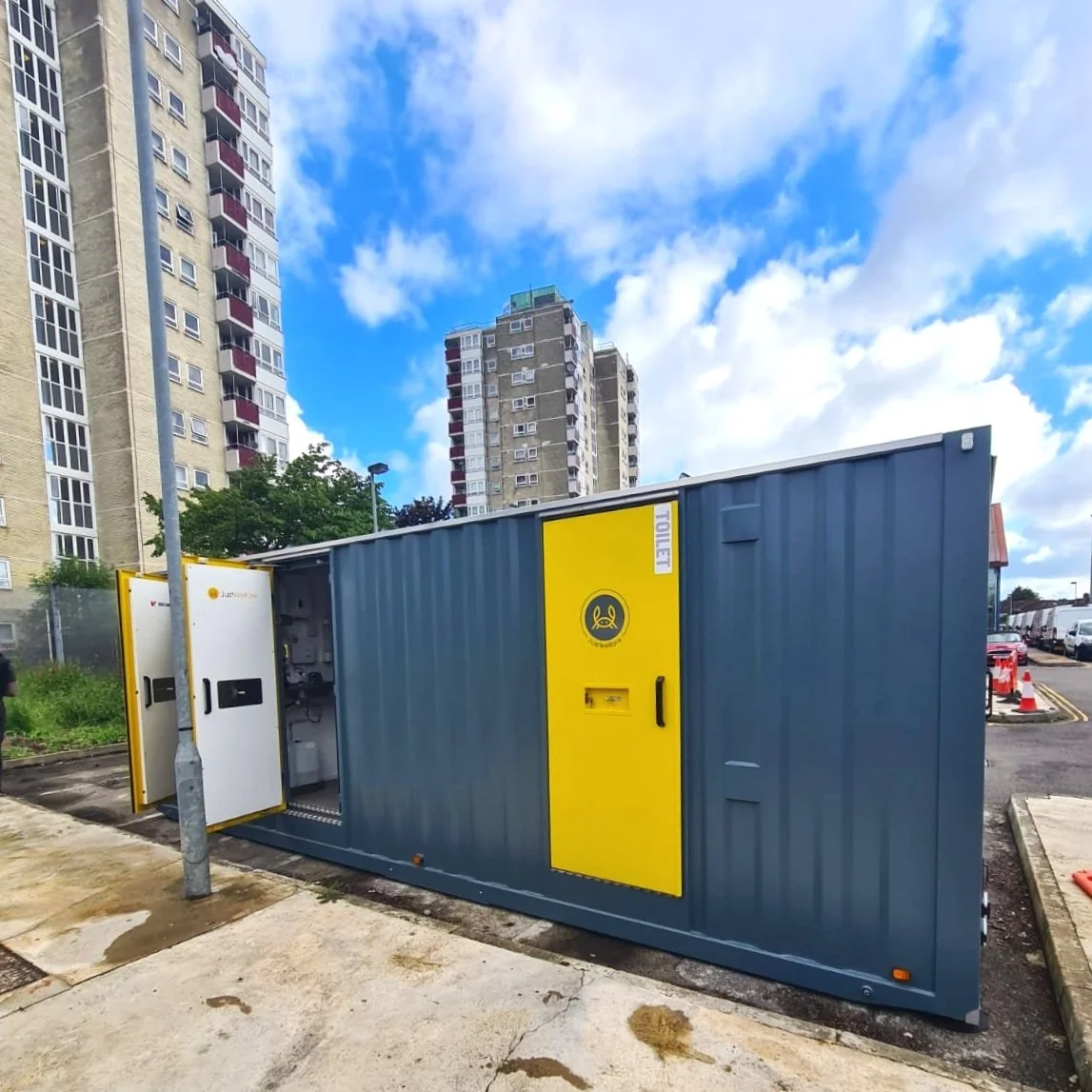 A blue and yellow portable electrical substation unit on a city street, with high-rise apartment buildings in the background under a partly cloudy sky.