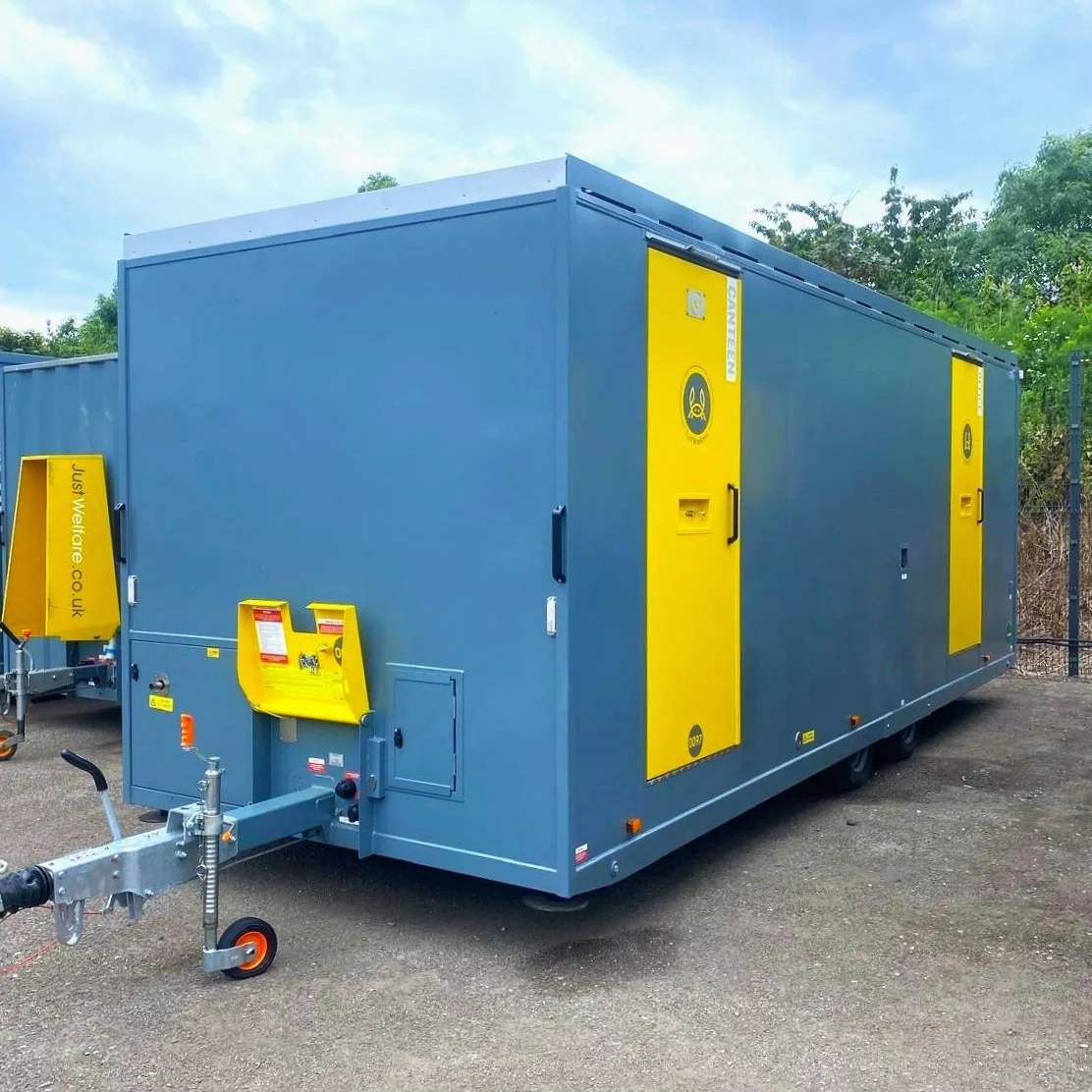 A large blue mobile quarantine or biohazard containment unit with yellow biohazard warning signs and doors, parked on a gravel surface with trees in the background.