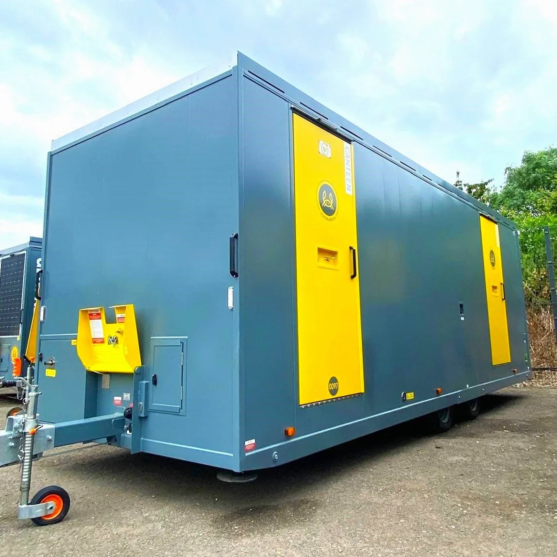A large gray generator with yellow doors and black and yellow Hazard labels, situated outdoors on a gravel lot, with some trees and a partly cloudy sky in the background.