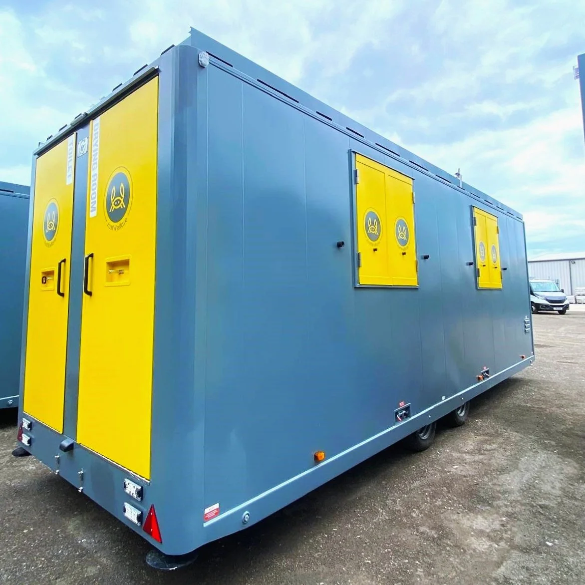 A large blue and yellow JUUL vaporizer trailer with JUUL logos, parked outdoors on a gravel lot under a cloudy sky.