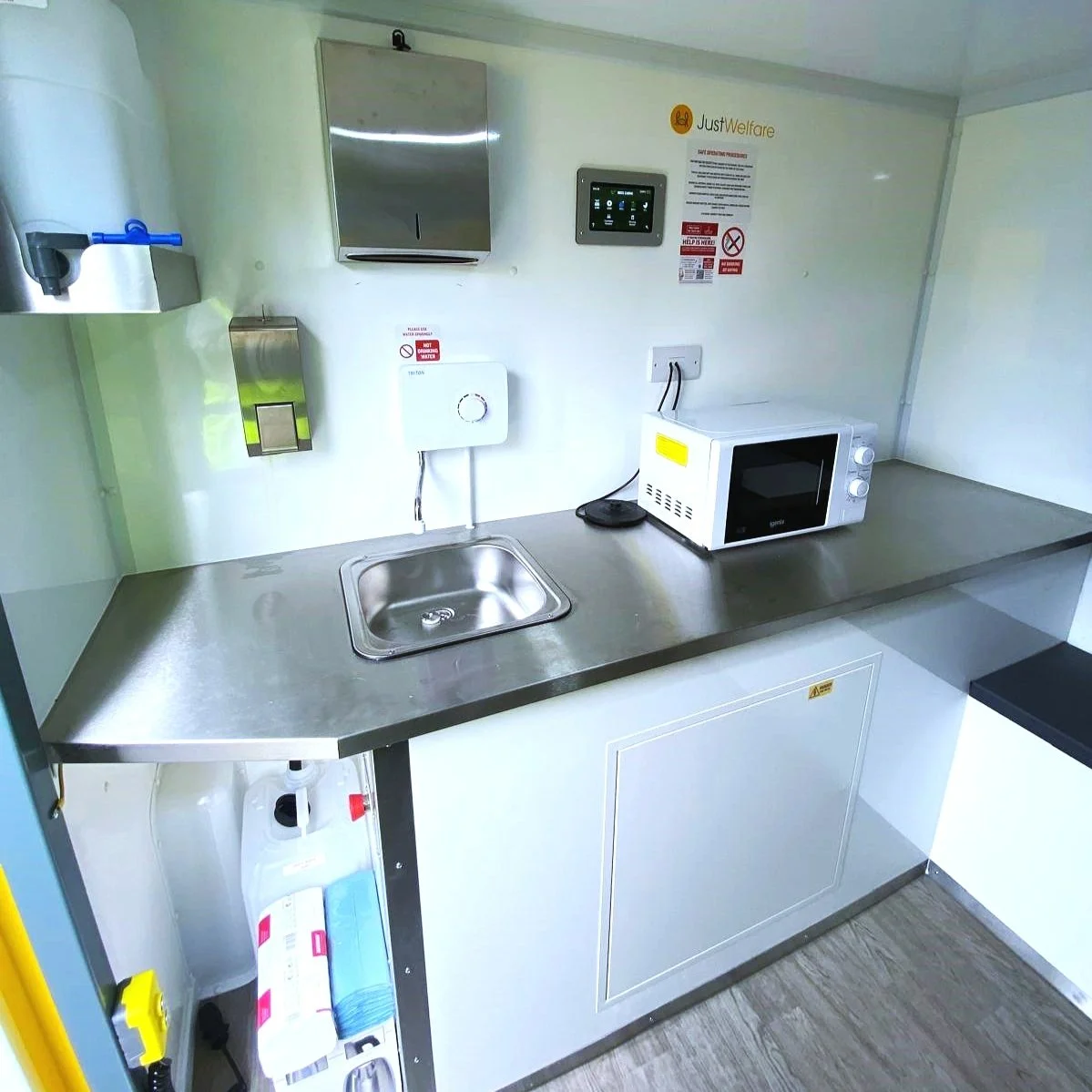A small kitchenette with a stainless steel countertop, a small sink, a microwave oven, and various wall-mounted equipment and signs.