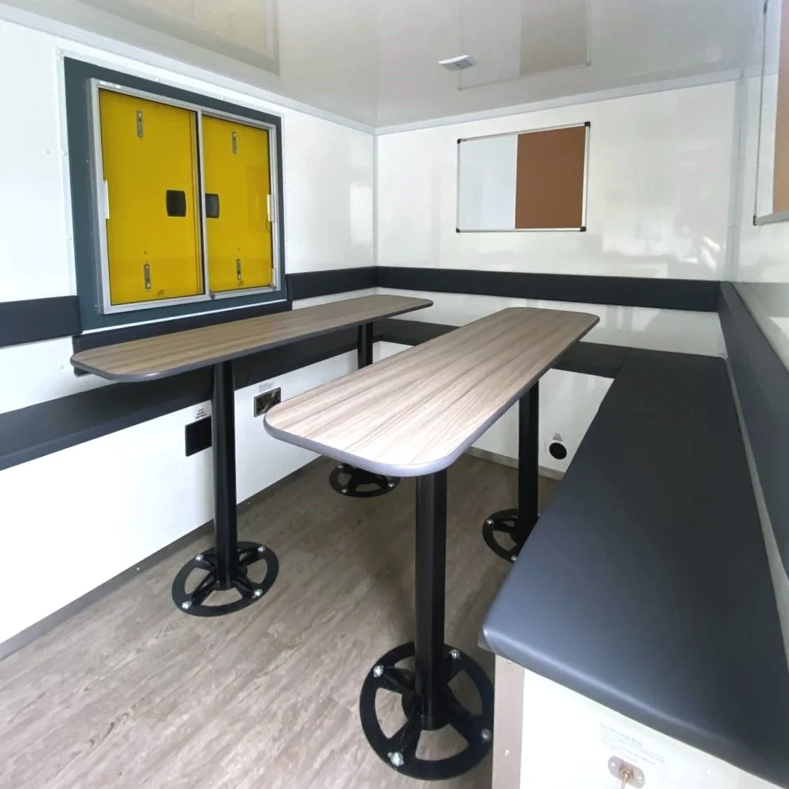 Small, enclosed room with two wooden tables, black corner seating, white walls with black trim, a yellow electrical panel, and a small window.