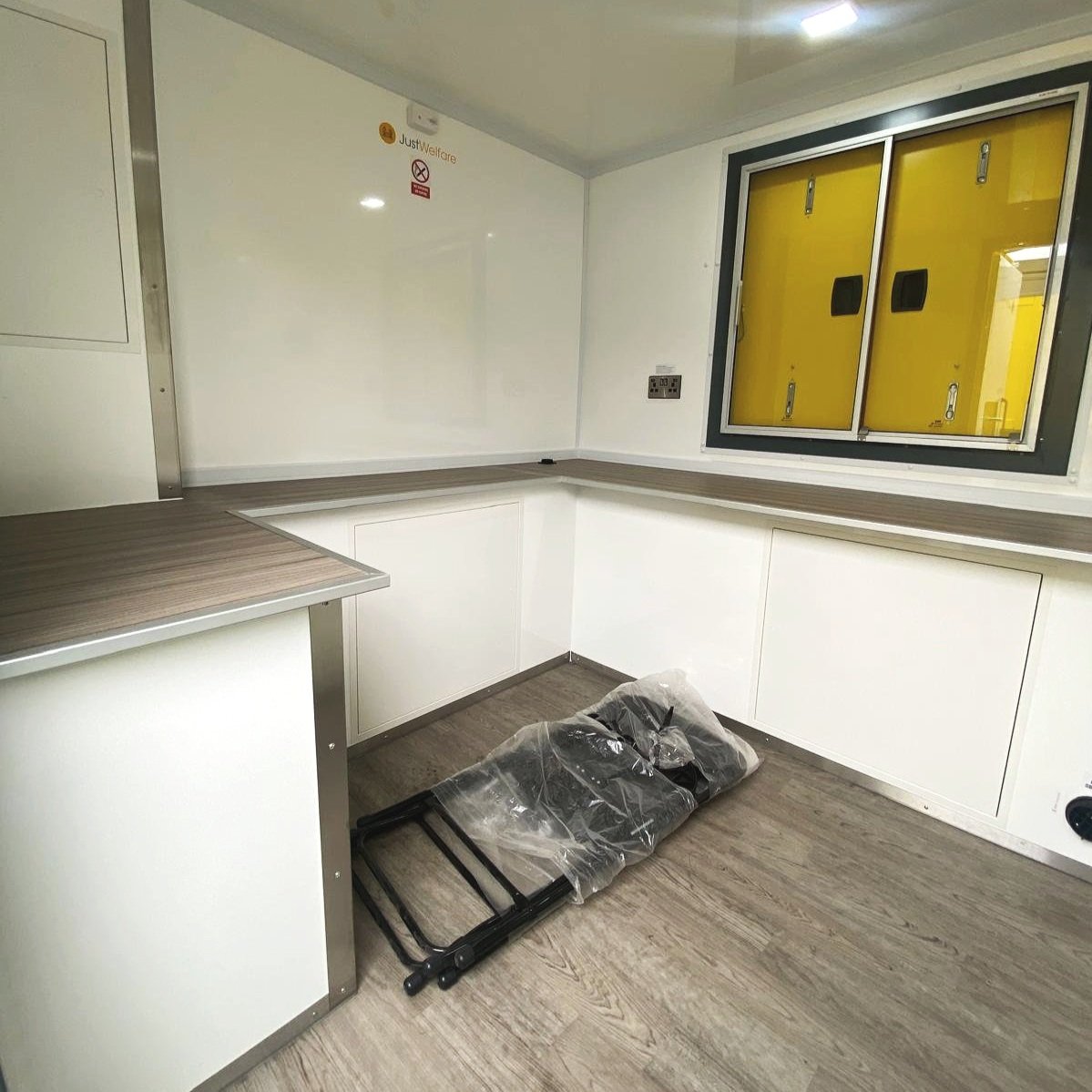 Empty mobile medical unit with a chair wrapped in plastic on the floor, yellow exterior visible through the window, and white walls with a small sign.