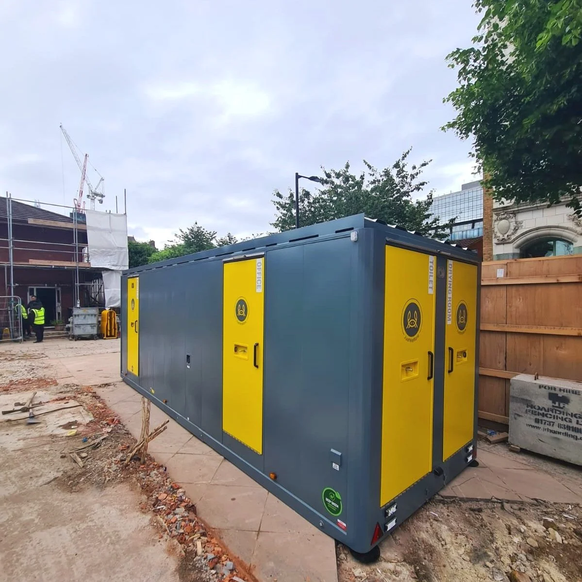 Portable toilet with yellow doors at a construction site, with construction workers and building materials in the background.