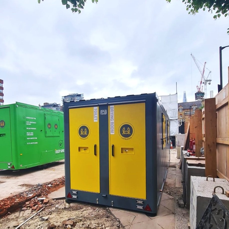 A yellow and black portable toilet at an outdoor construction site, with a green generator in the background and construction materials around.