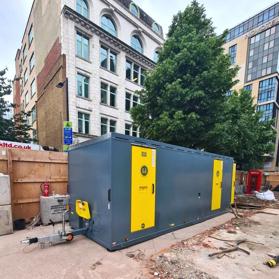 A large gray and yellow portable generator on a construction site in an urban area with multi-story buildings, a tree, and construction materials around.