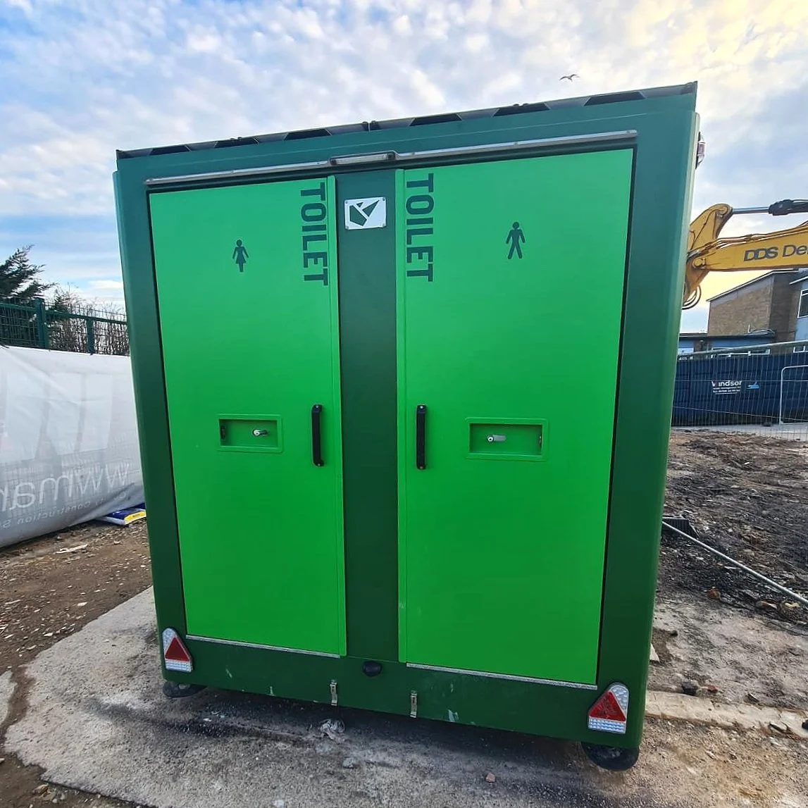 Green public toilet with women's symbols on the doors, set against a partly cloudy sky and construction site background.