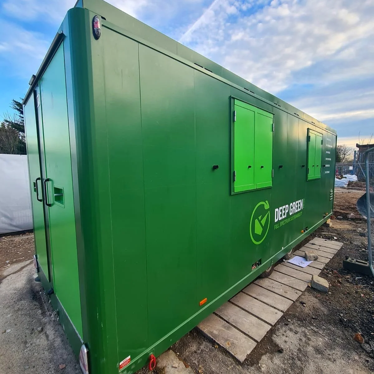 A large green container with the words 'Deep Green' and 'Full Spectrum Sustainability' printed on it, situated outdoors on a construction site with a partially cloudy sky in the background.