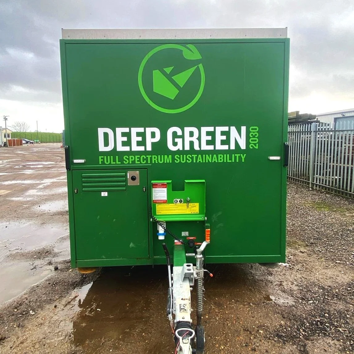 A green trailer with the words "Deep Green 2030 Full Spectrum Sustainability" and a recycling symbol at the top. The trailer is on a muddy, wet ground outdoors with a cloudy sky.