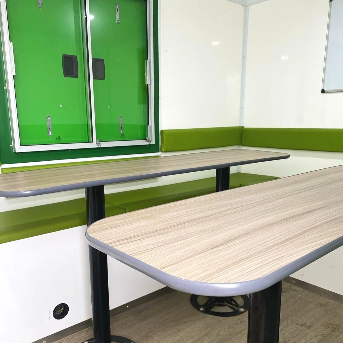 Empty classroom with two light wood tables, green benches along the wall, and a green storage cabinet with sliding doors on the wall.