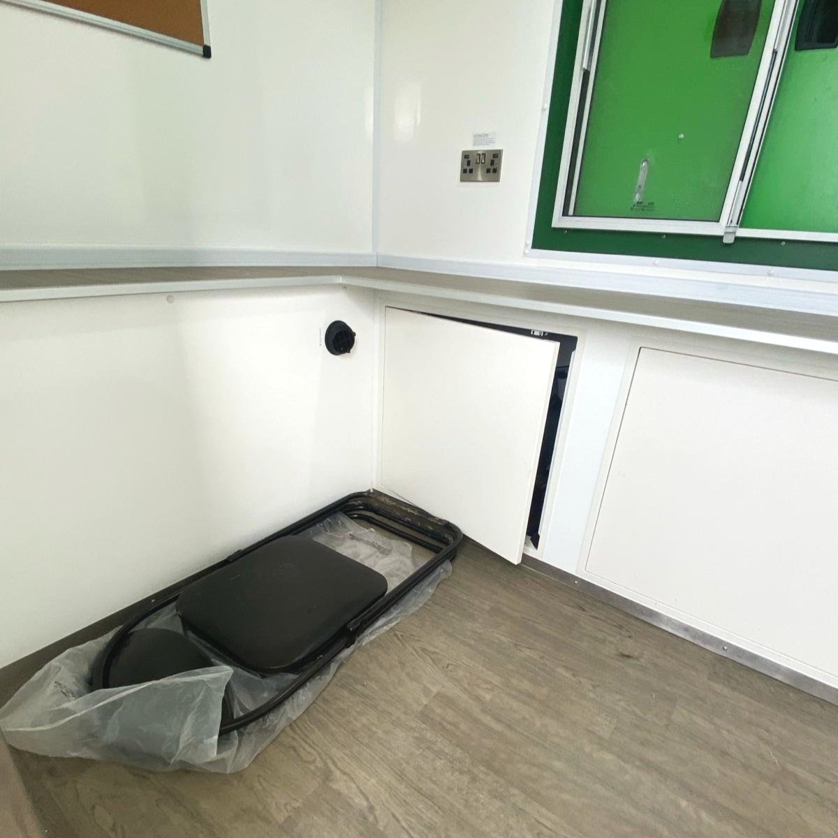 Empty white medical or laboratory room with closed cabinets, a green window with black panels, and a black object in plastic at the corner of the floor.