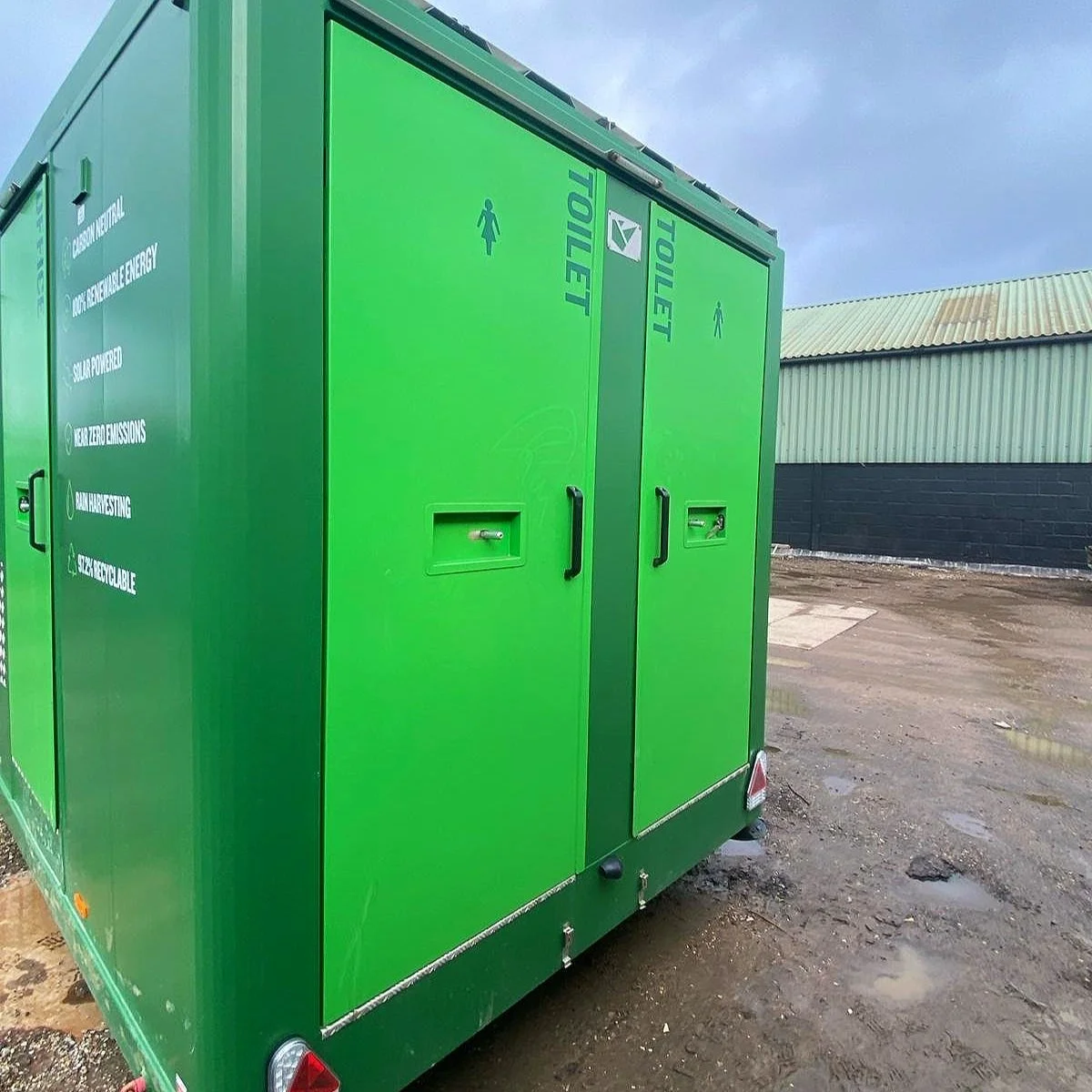 Green mobile restroom trailer with designated men's and women's toilets and accessible features, parked outdoors on a dirt surface near a building with a metal roof.