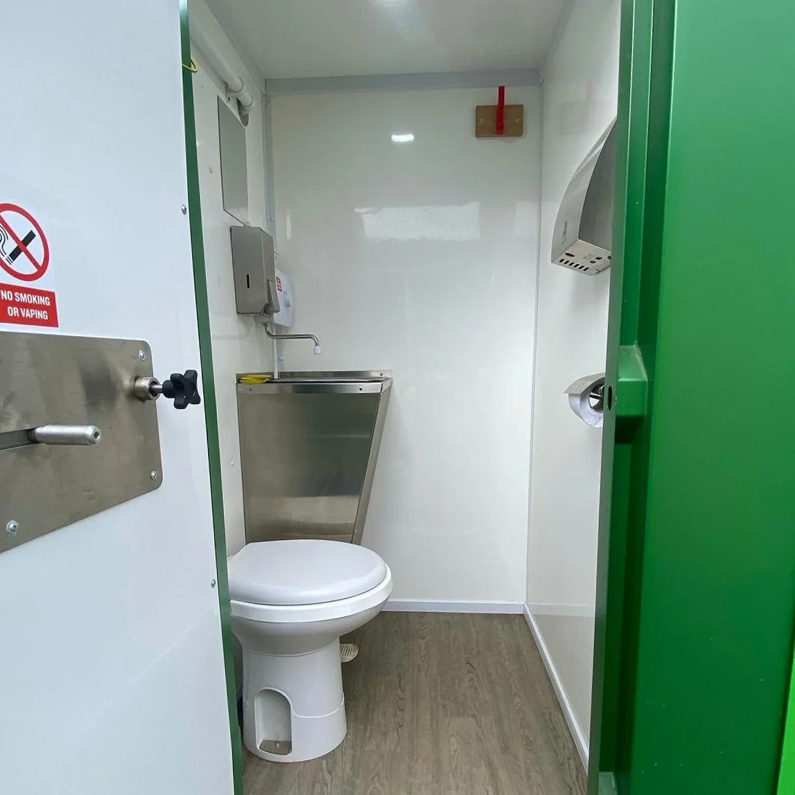 Small clean restroom with a white toilet, stainless steel sink, paper towel dispenser, and a vent hood. No smoking or vaping sign on the door.