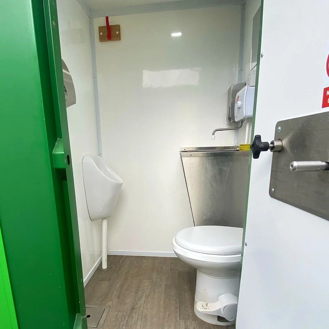Small men's restroom with a white toilet, a urinal on the left wall, and a stainless steel sink at the back, with a soap dispenser on the wall above the sink.
