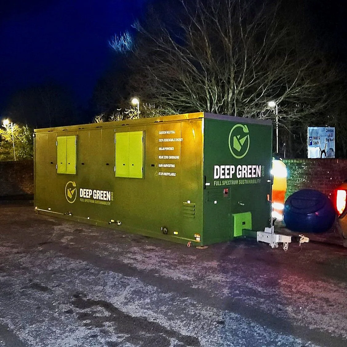 A green mobile unit with the logo and name 'Deep Green' parked outdoors at night. The unit features several windows with closed shutters and lights illuminating the scene. A black tire is attached to the front, and the background includes trees and s