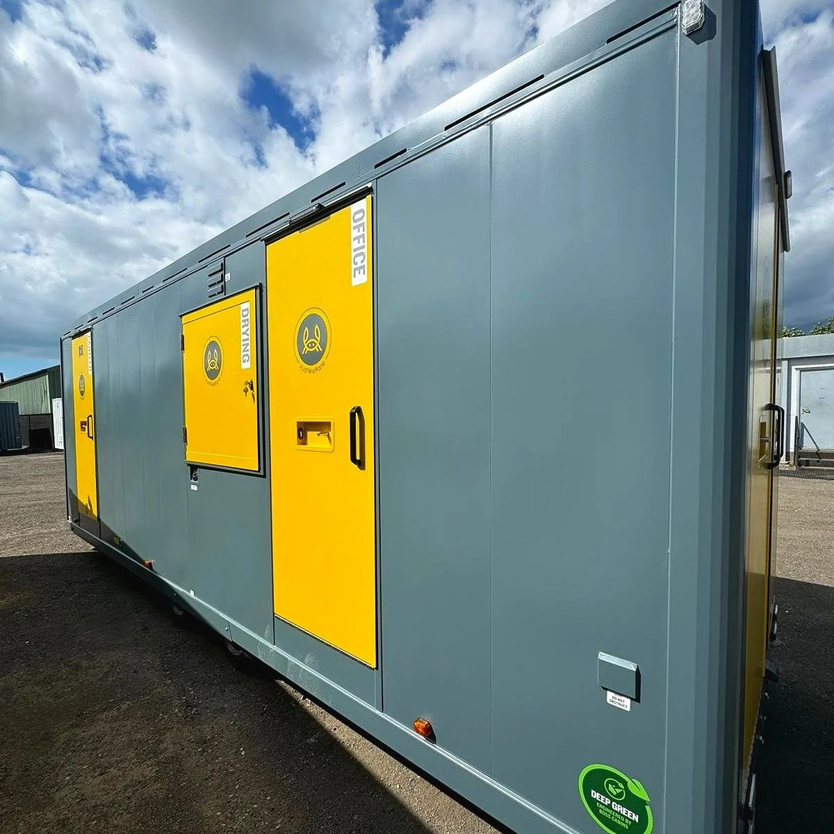 A large gray utility trailer with yellow doors labeled for office and driving purposes, parked on a gravel lot under a partly cloudy sky. There is a green sticker at the lower right corner indicating eco-friendliness.