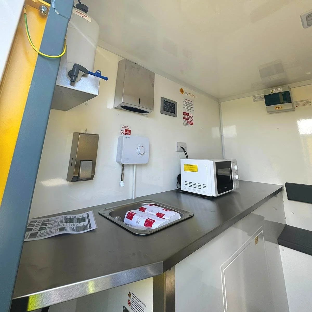 Small kitchenette with stainless steel countertop, microwave, disposable cups, and wall-mounted soap dispenser, electrical outlets, and warning signs.