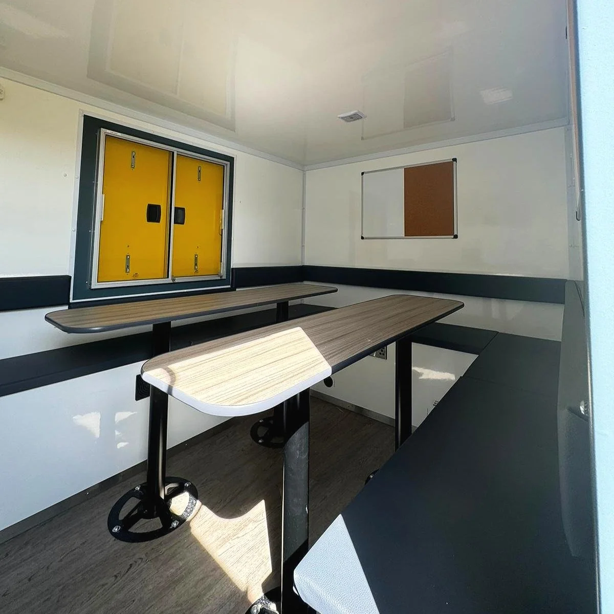 Inside a small, empty room with folding yellow electrical panels on the window, a whiteboard with a brown bulletin board, and a set of tables and benches for meetings or work.
