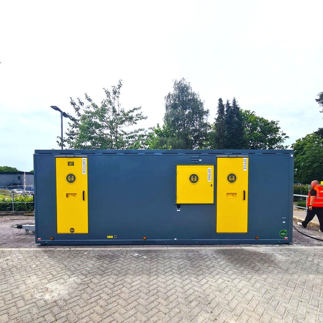 A dark gray container with incorporated yellow doors and panels labeled for caution, warning of biological or hazardous materials, with a person in a red safety vest walking nearby in an outdoor parking lot.