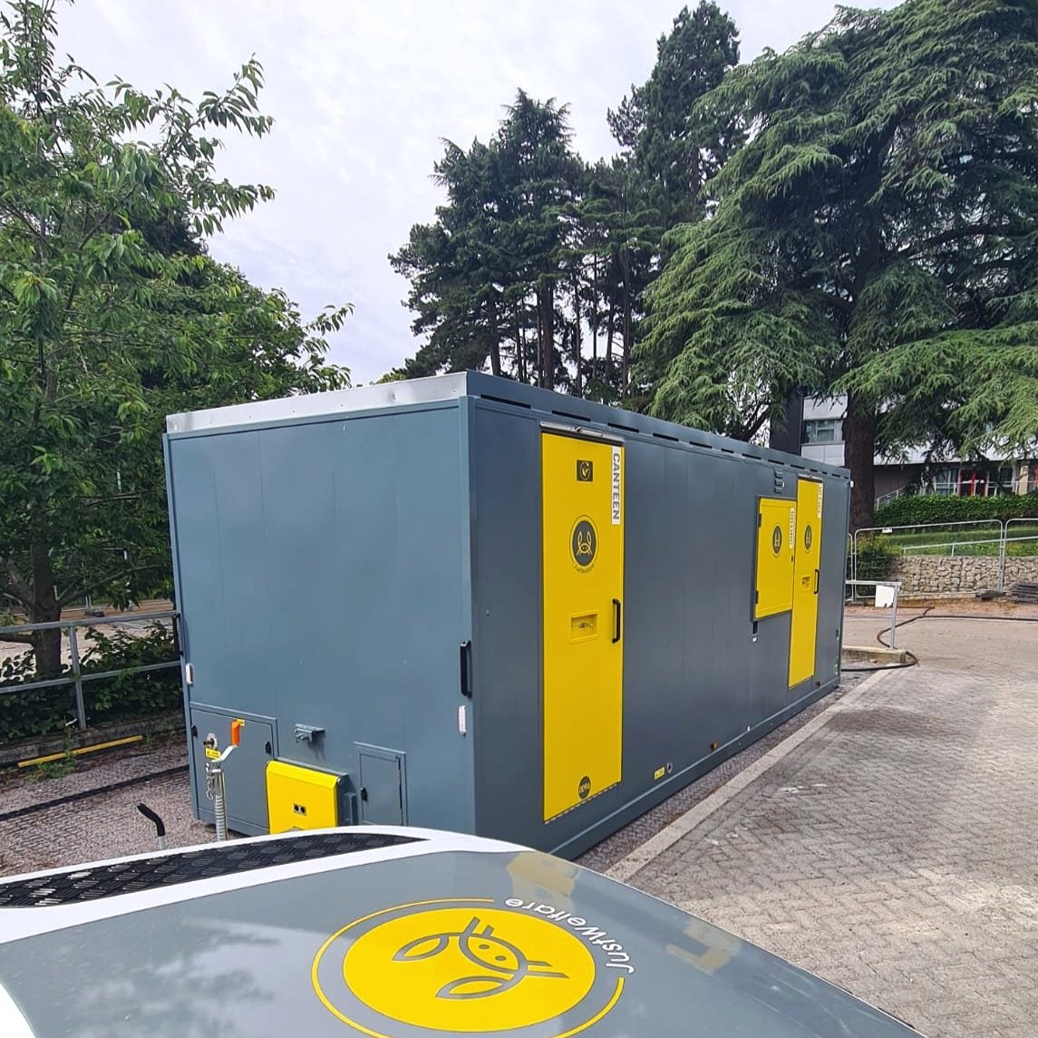 A mobile electrical utility unit with gray and yellow panels, situated on a paved lot with trees and a building in the background.