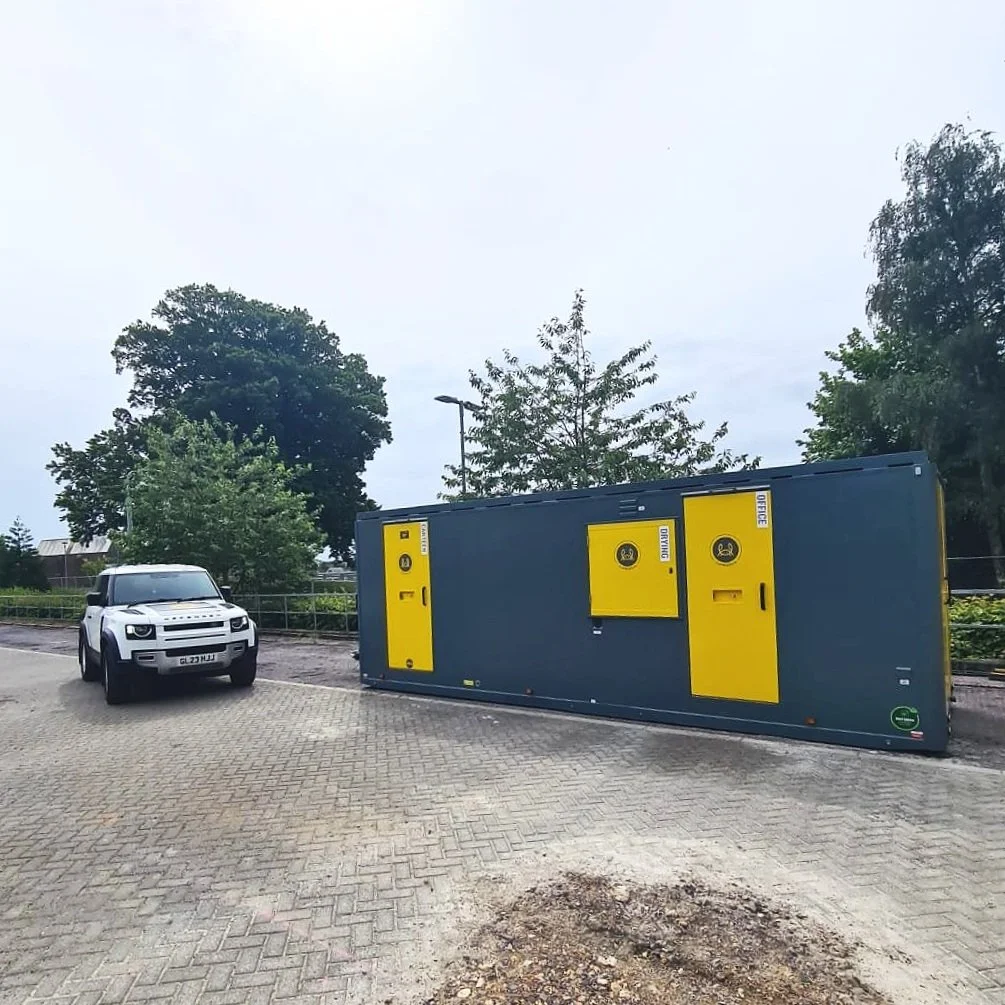 A white SUV parked next to a blue and yellow USPS mailbox unit on a paved area, with trees and an overcast sky in the background.
