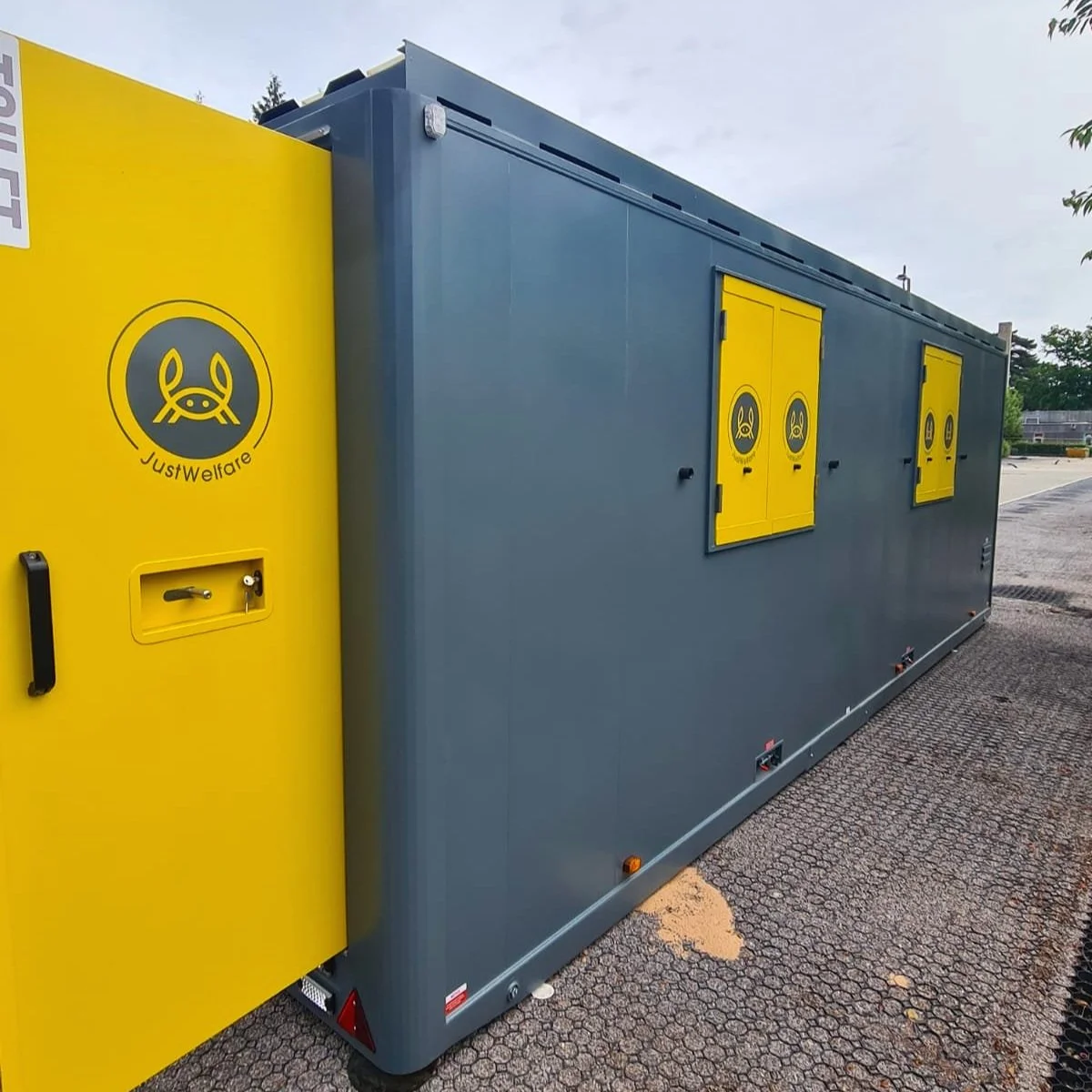 A large rectangular mobile unit with a gray body and yellow doors and panels. The yellow section has a logo with a pig face and the words 'JustWelfare.' The unit has multiple vents, windows, and access doors, likely used for animal welfare or veterin
