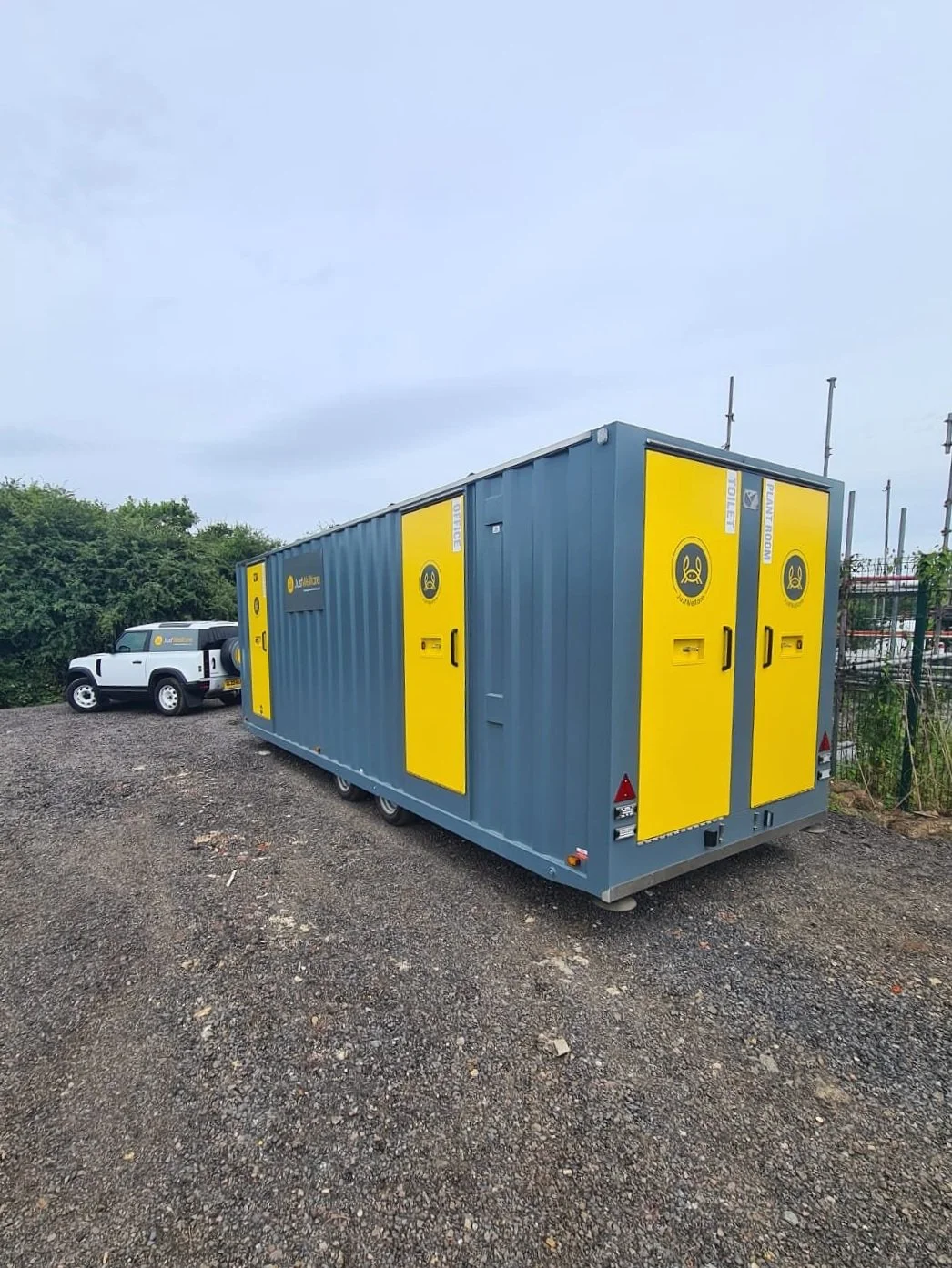 A mobile dental clinic in a blue and yellow container with a logo and signage, parked on gravel near some green bushes, with a white off-road vehicle behind it.