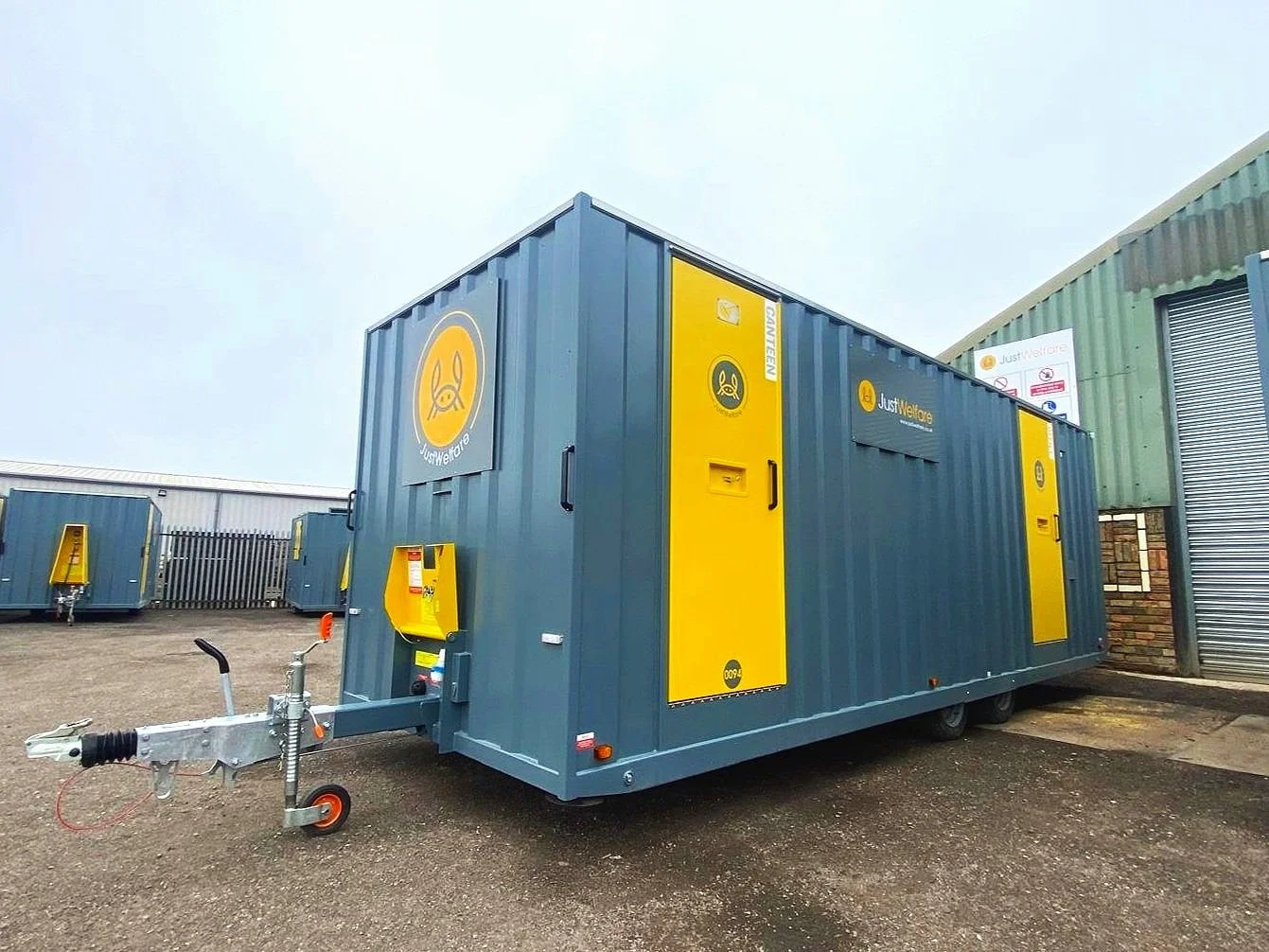 A large blue and yellow portable restroom trailer marked with 'Just Welfare' logo and branding, parked on a gravel lot near industrial buildings.