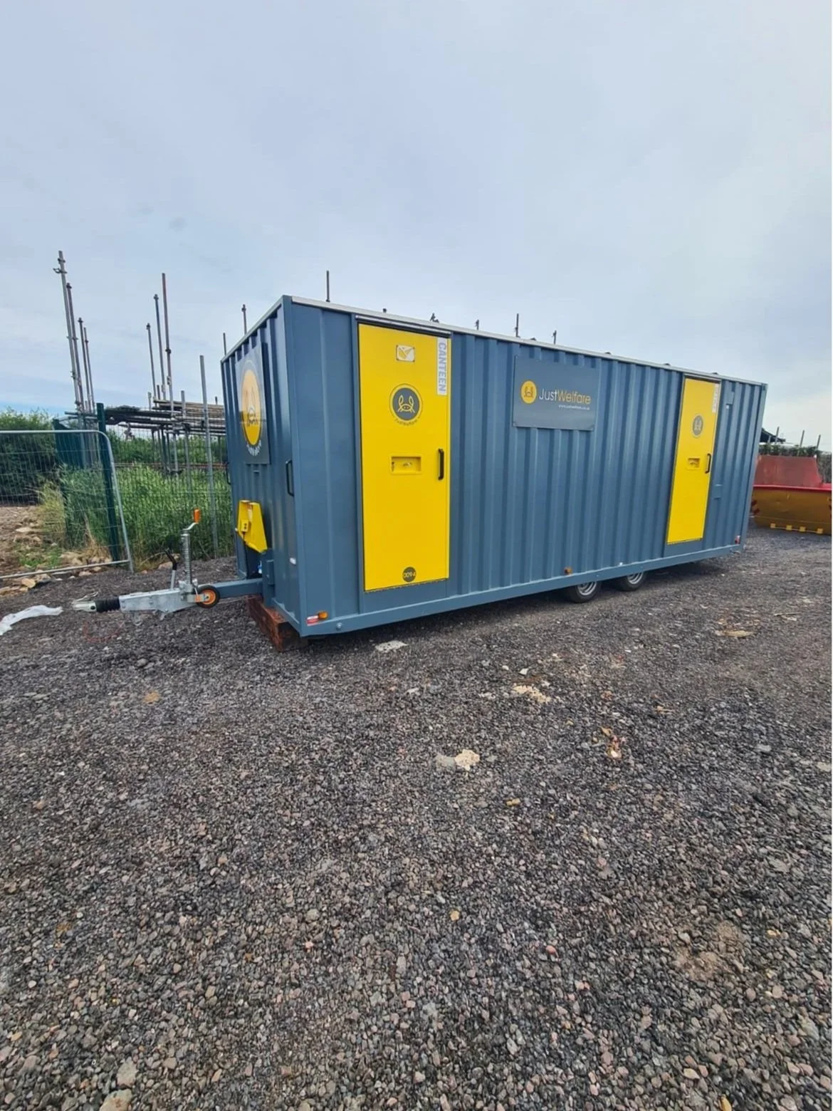 A large blue container with yellow doors labeled 'JustWelfare' and 'Canteen' sits on gravel ground with construction equipment and fencing in the background.
