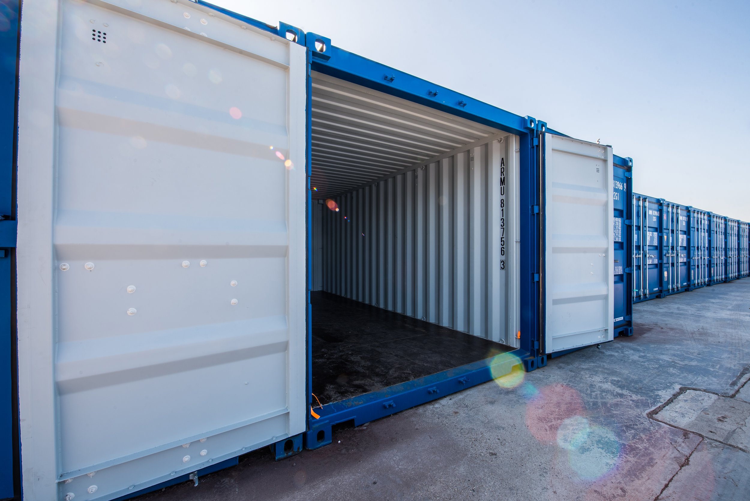 Open blue shipping container on a concrete surface with more containers stacked in the background under clear sky.