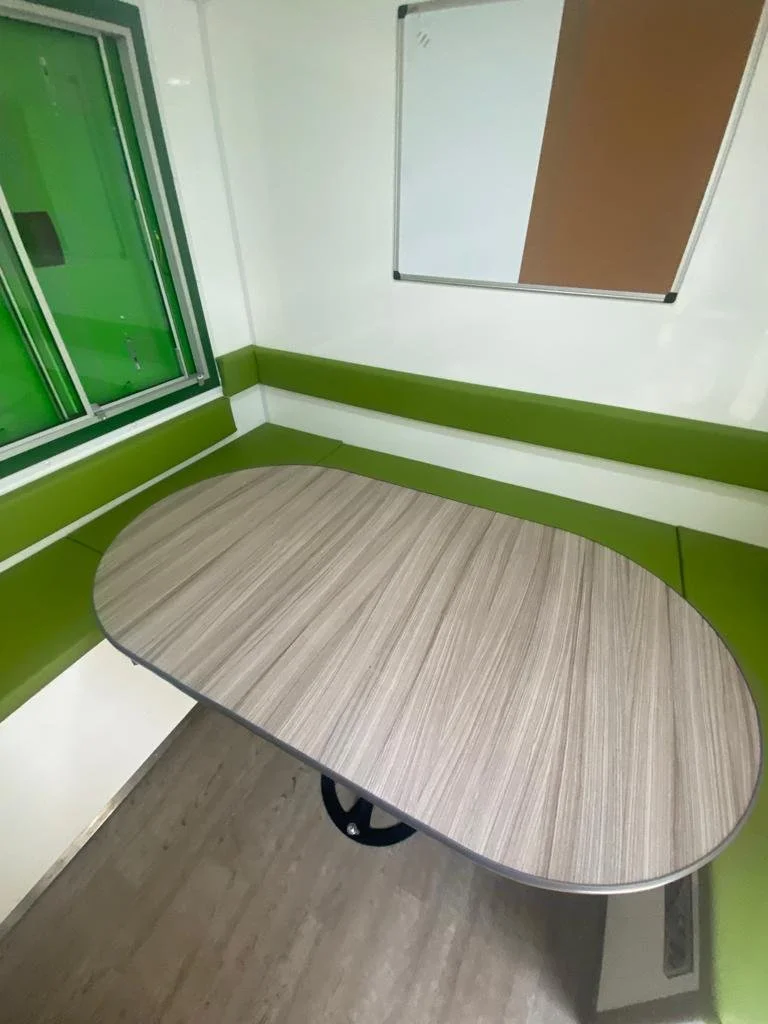 A small room with a green semi-circular bench, a wood-patterned table with a wheel underneath, a whiteboard, and a window with green blinds.