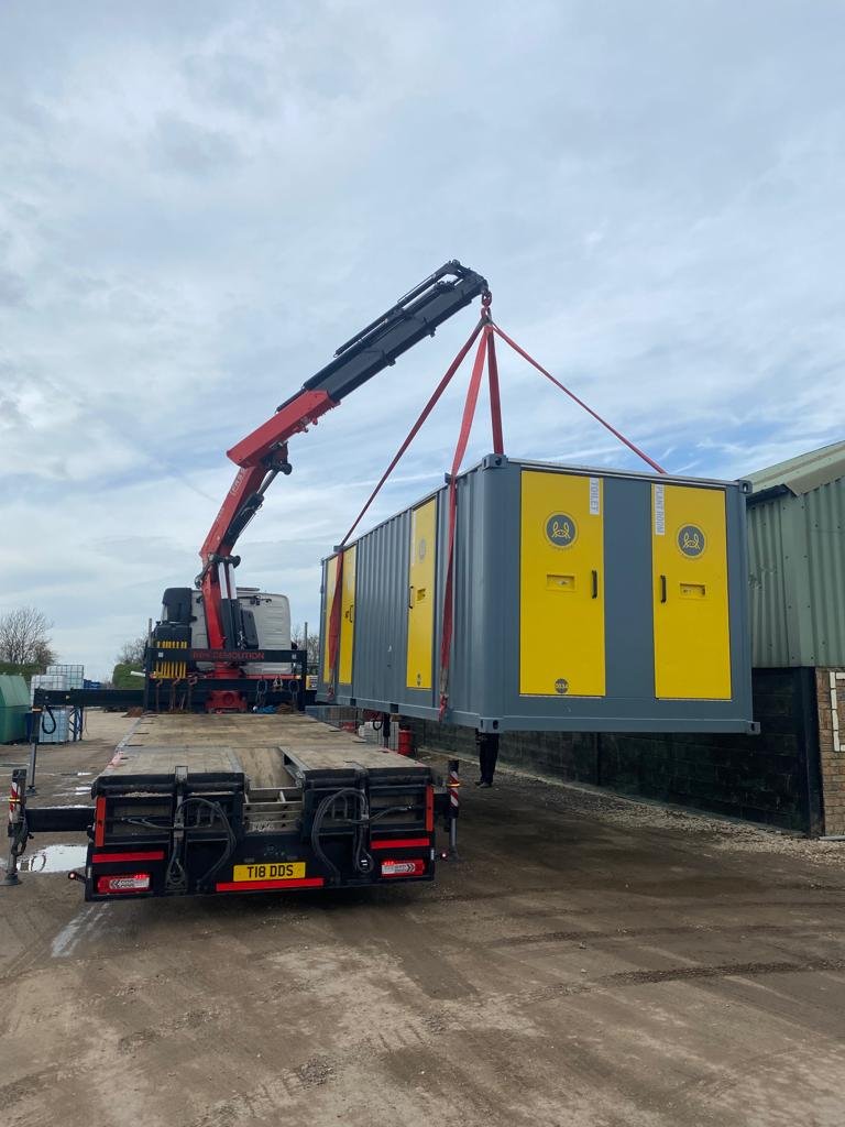 A construction crane lifting a large gray and yellow electrical cabinet onto a trailer.