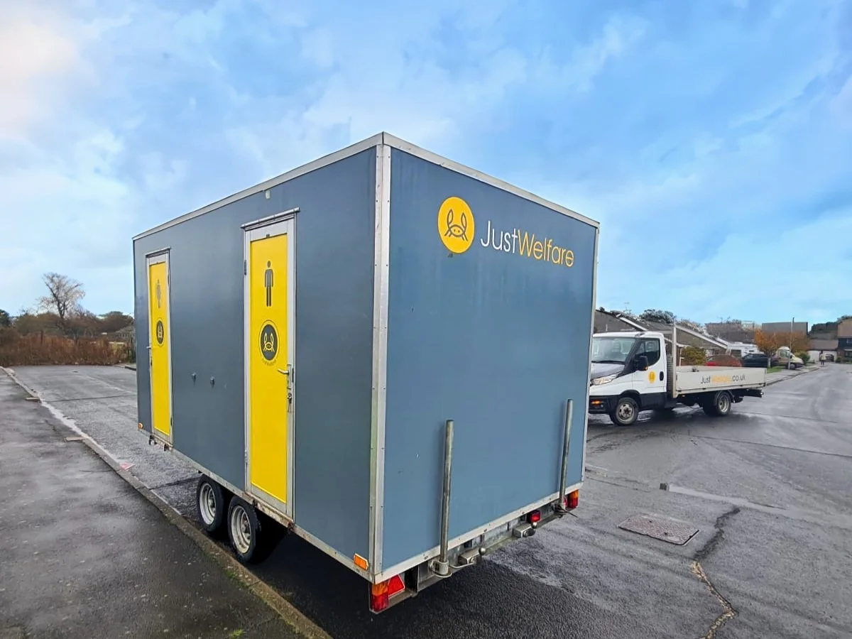 A mobile restroom trailer with three yellow doors, each displaying icons for male, female, and waste. The trailer has a blue exterior with a logo for "JustWelfare" and is parked in an outdoor lot.