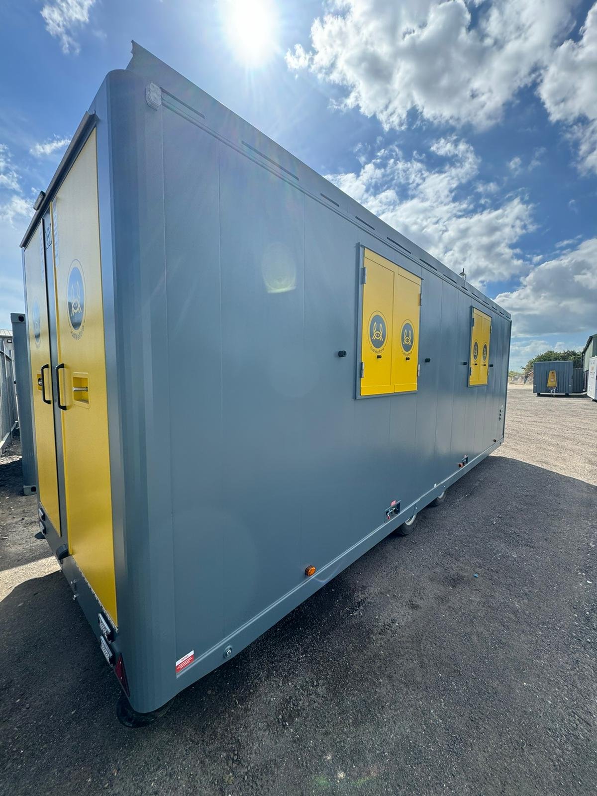 Large gray mobile unit with yellow doors featuring logos, situated outdoors on a gravel surface under partly cloudy sky.