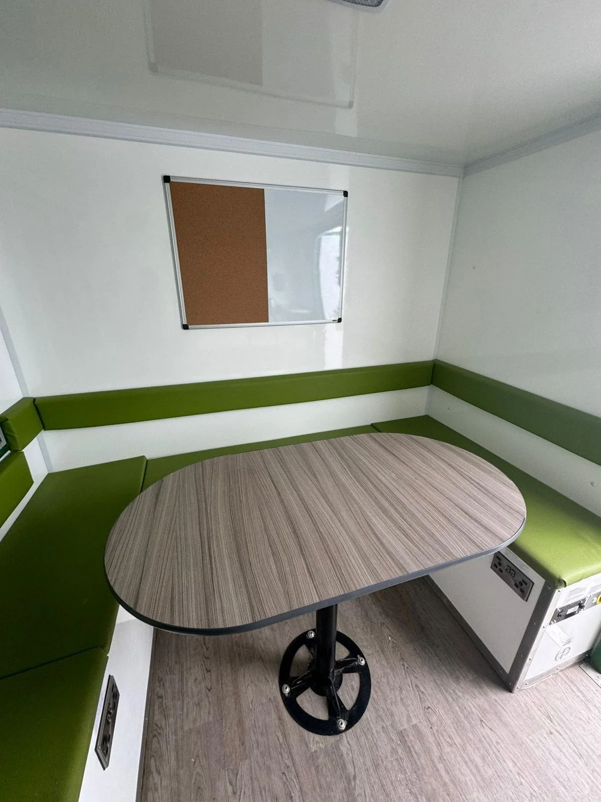 Small dining area with L-shaped green cushioned seating and a wooden oval table, a mirror on the white wall, and a wood floor.