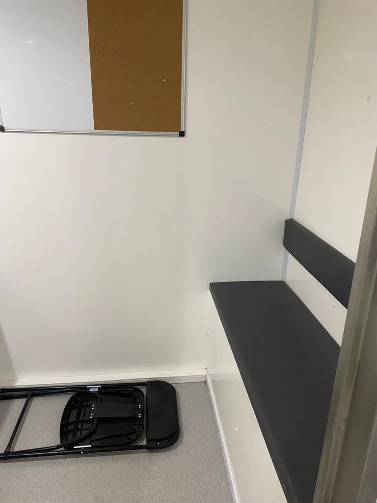 Empty small room with a black bench, a cork bulletin board, a folding dolly, and plain white walls.