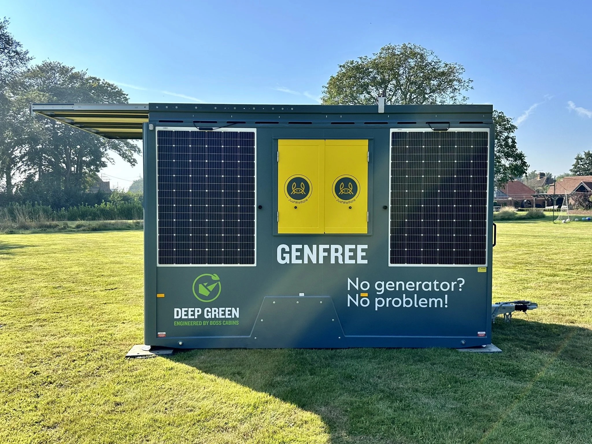 A green portable power station with solar panels on the sides, placed on grass in an open field with trees and houses in the background. The station has a yellow door with logos, and text that reads 'GENFREE', 'No generator? No problem!', and 'DEEP G