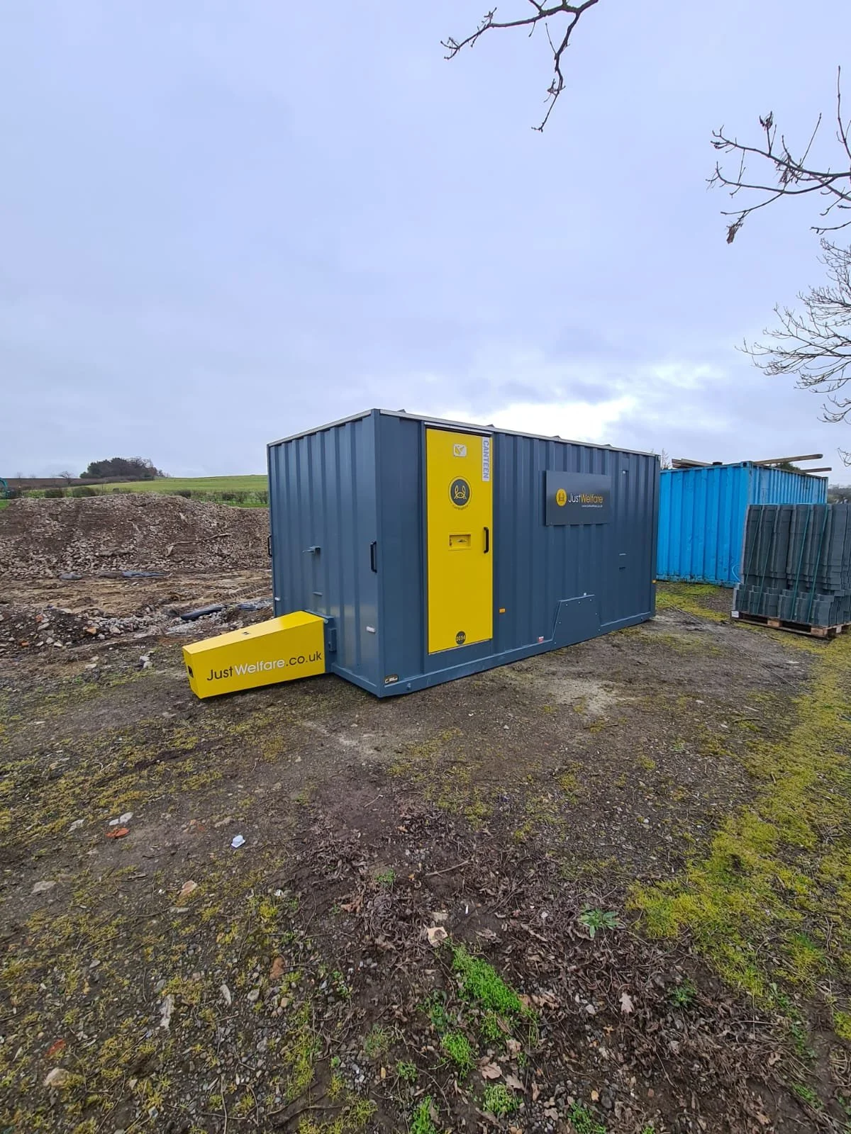 A portable welfare unit with blue and yellow colors on a dirt and grass ground, with a blue sky above and some trees in the background.