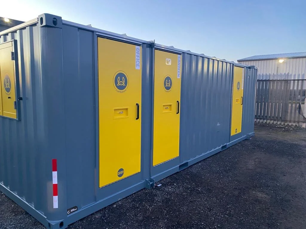 A blue shipping container with yellow safety doors and logos, set on gravel outdoors under a blue sky.