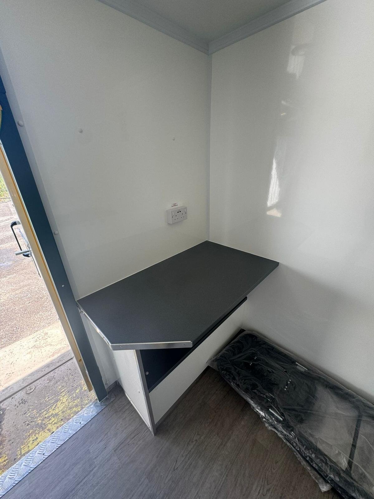 An empty room corner with a black countertop attached to the wall, a power outlet, and a black pad or mat on the floor wrapped in plastic.