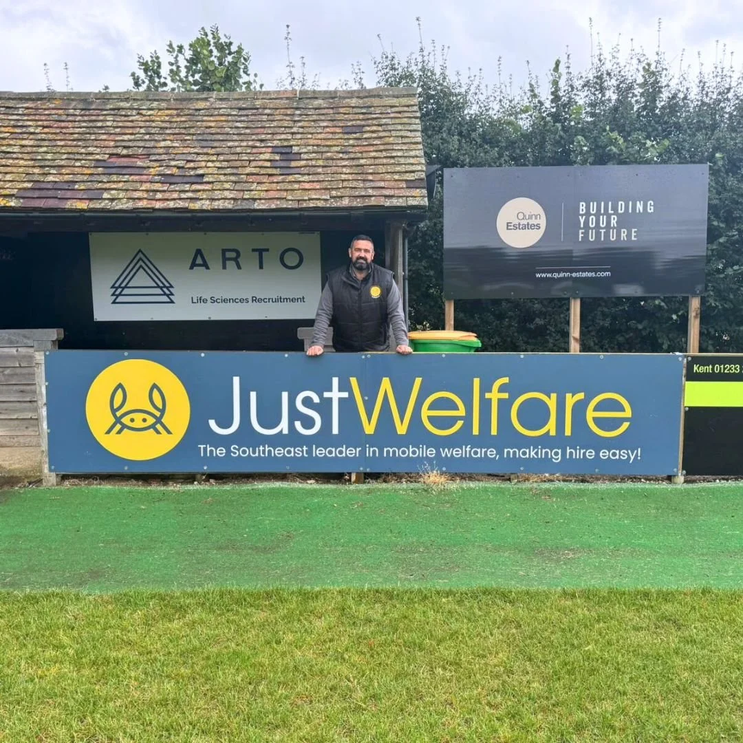 Person standing behind a large blue and yellow sign that reads "JustWelfare" with a tagline about mobile welfare. The sign features a yellow logo of a crab and the text "The Southeast leader in mobile welfare, making hire easy!" Behind the person are additional signs for Arto Life Sciences Recruitment and Quinn Estates, with trees and a cloudy sky above.