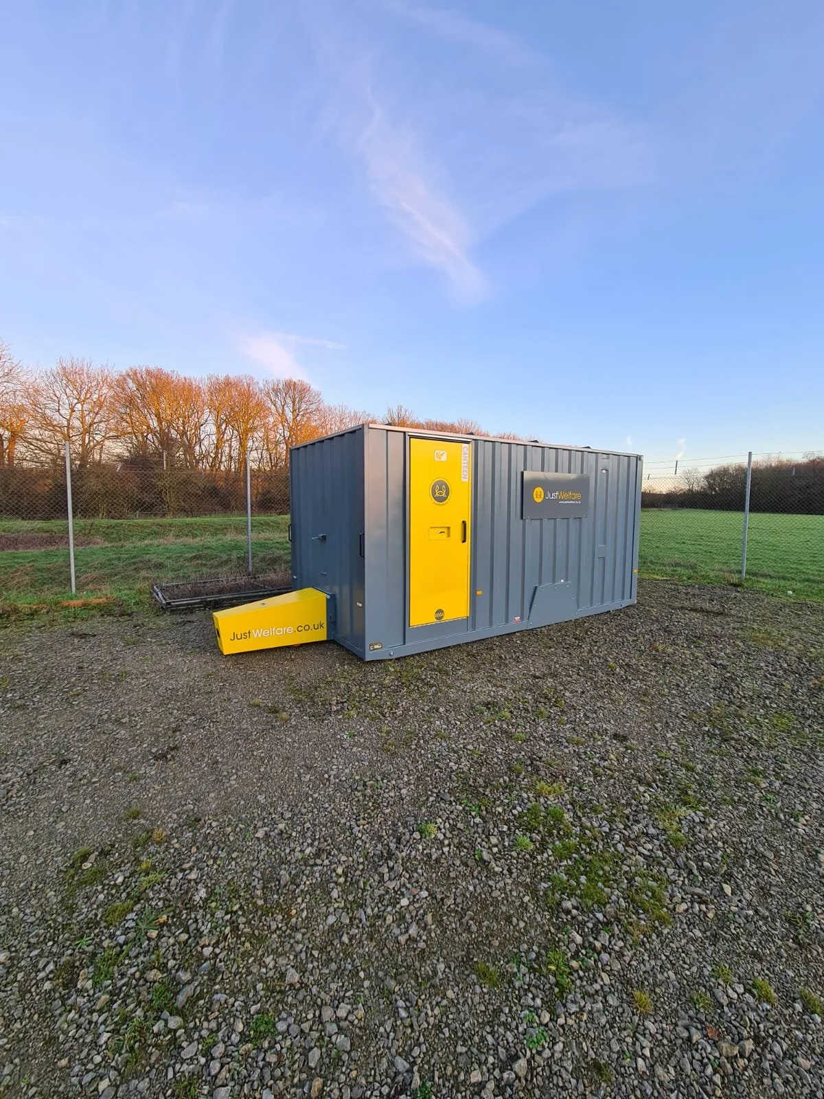 A portable welfare facility container painted in grey with yellow accents, placed on a gravel surface next to a chain-link fence and open field, under a clear blue sky.