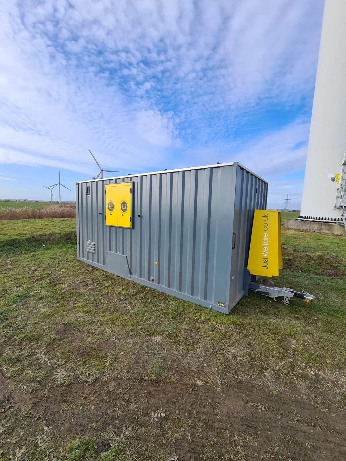 A large gray metal container with yellow doors and yellow signage, situated on grass outdoors with wind turbines and power lines in the background under a partly cloudy sky.