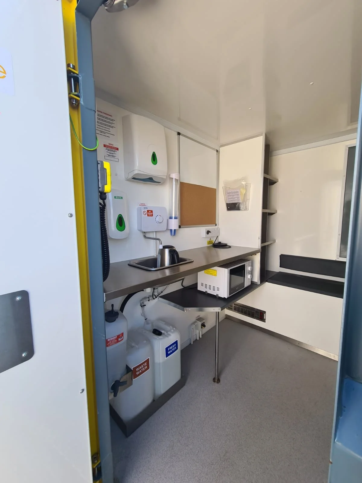 Small medical or laboratory room with a stainless steel countertop, microwave, and various dispensers on the wall, including hand sanitizer and soap, with a small sink and water containers underneath.