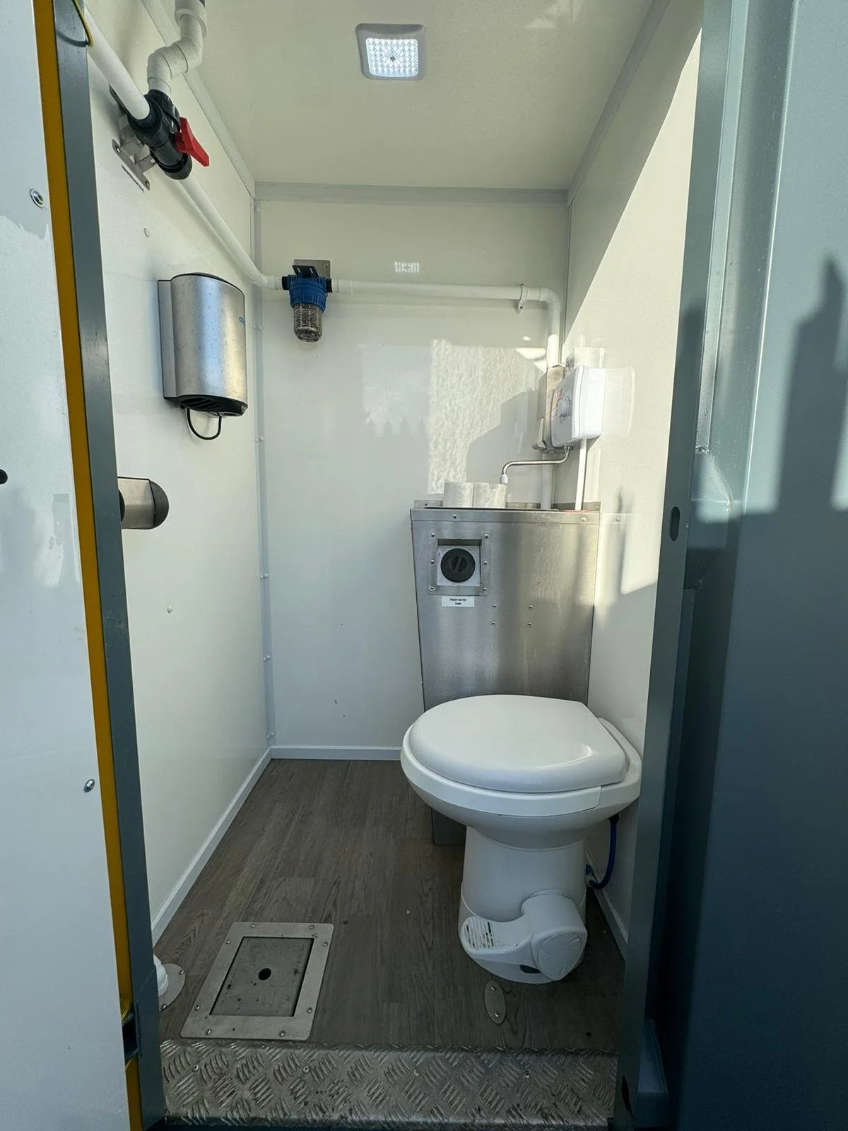 Small, white bathroom with a toilet, metal waste bin, and plumbing fixtures on the wall. The floor is gray wood-covered, with a metallic floor drain in front of the toilet.