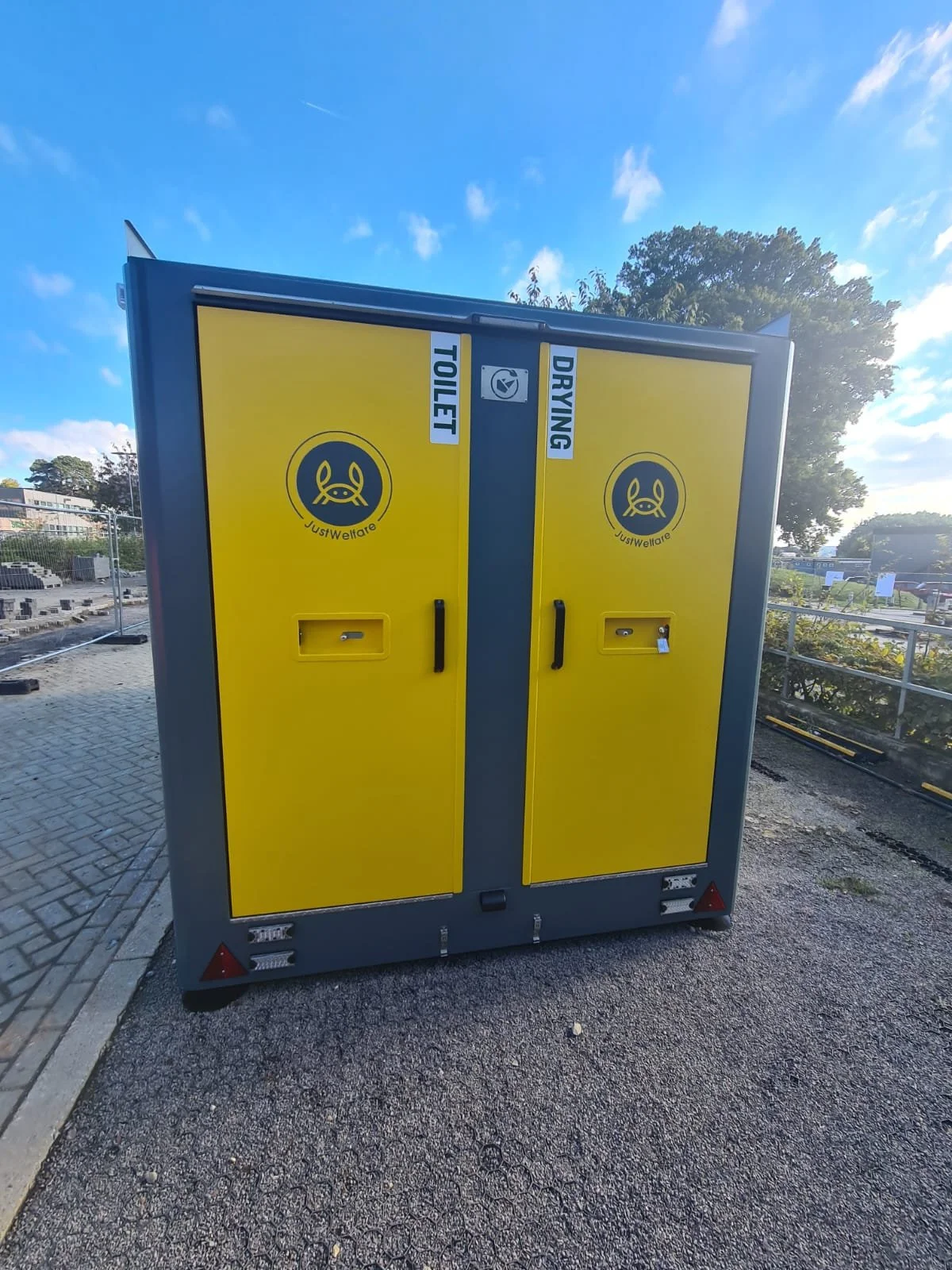 Yellow and gray public toilet and drying station with signs labeled 'Toilet' and 'Drying' near trees and blue sky.