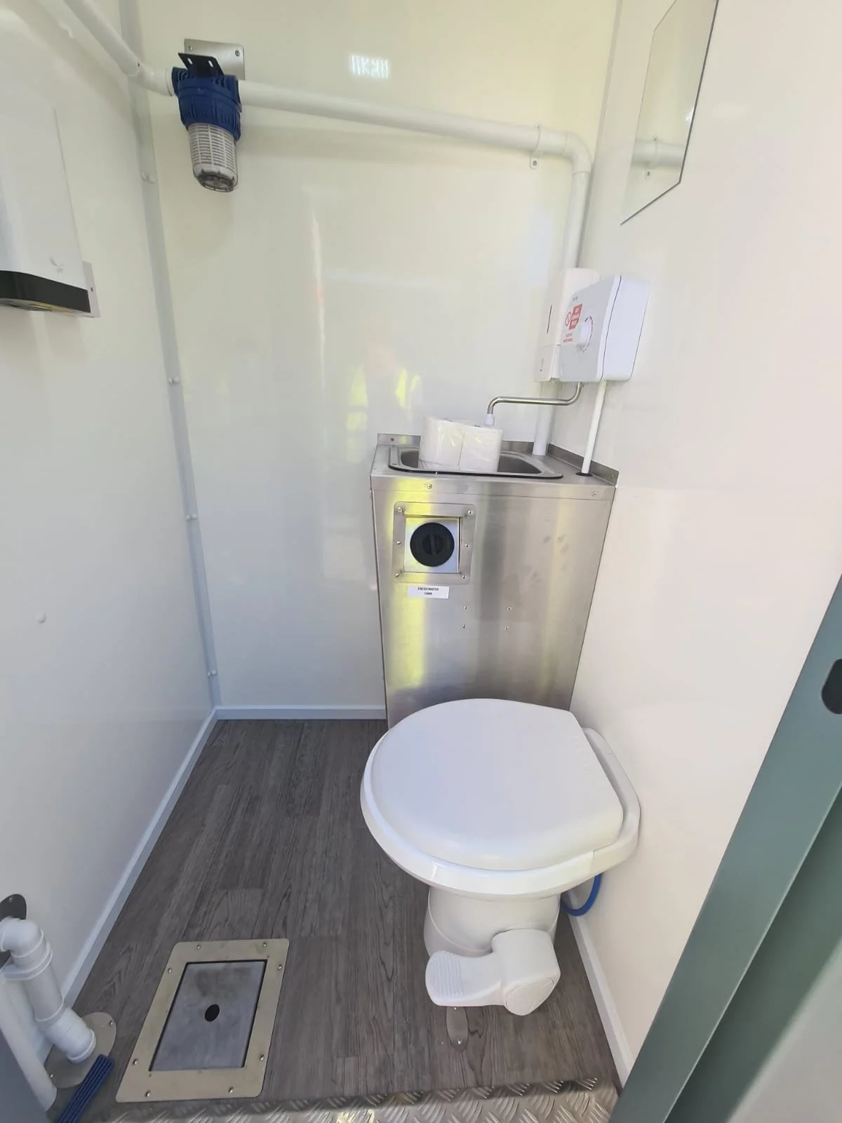 Small bathroom with a white toilet, a metal sink with a paper towel roll, and plumbing pipes, including a large plumbing vent pipe and an electrical or plumbing box, all in a clean, minimalist setting.