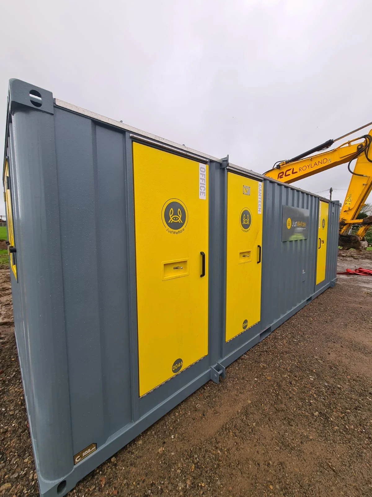 A large gray and yellow portable electrical box with multiple doors, labeled with signs such as "Office" and "Maintenance," sitting on dirt ground with construction equipment visible in the background.
