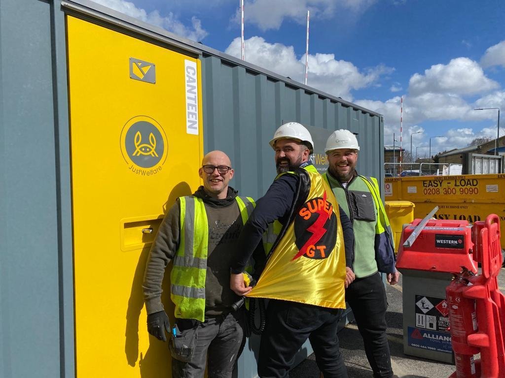Three men wearing safety vests and hard hats standing outdoors in front of a large gray container with a yellow door and a caution sign. They are smiling and looking at the camera.