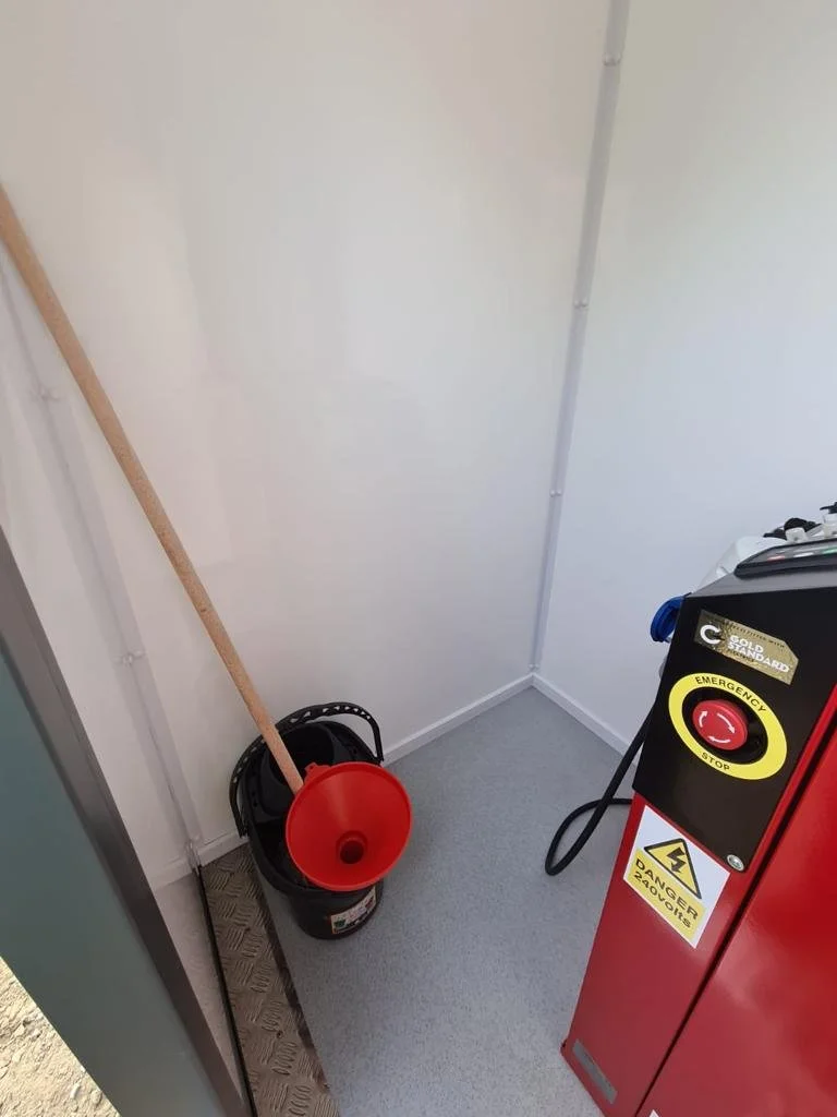 A small room with a mop bucket, mop, and a fire alarm or emergency stop button on a red panel.